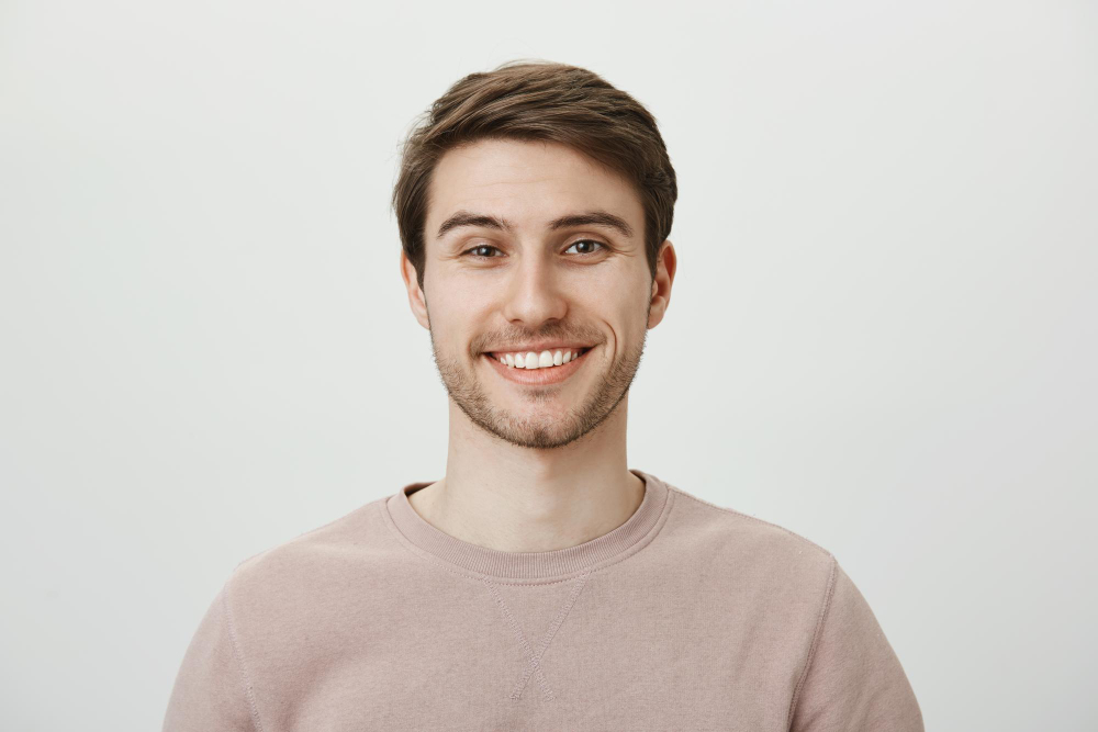 confident-attractive-caucasian-guy-beige-pullon-smiling-broadly-while-standing-against-gray